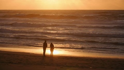 Couple Walking By The Ocean At Sunset, Lens Flare. SLOW NOTION. Unrecognizable man and woman romantic family silhouettes enjoying evening stroll by the stormy ocean tide. 