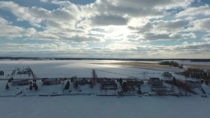 Drone footage panning around some beach houses on the side of The Ottawa River in the winter on a sunny and cloudy day.
