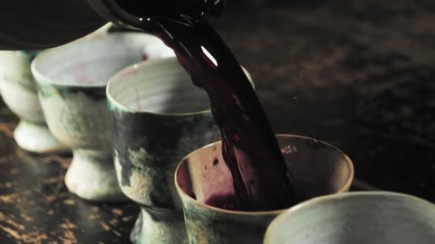 Slow Motion of red wine being poured from an ancient wine jug into a row of colorful wine goblets.