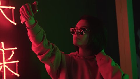Trendy cute girl blogger in sunglasses taking selfie photo or video using smartphone. Attractive brunette young woman looks at herself with mobile phone and touching her short hair. Red neon light