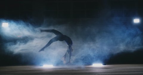 In slow motion in the smoke girl acrobat performs several back flips and coups in the air.