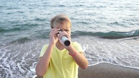 Closeup view of cute happy young kid standing at summer beach and looking through vintage old hendheld telescope at something interesting far away in distance. Real time 4k video footage.