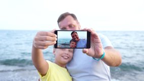 Cute family of adult bearded father and young handsome son taking selfie at seaside during summer vacations at hotel resort. People using digital camera of smart phone. Real time 4k video footage.