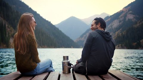 Beautiful happy real romantic couple of match man and woman friends in love on first date in nature having fun smiling or laughing give a high five and sitting on wooden bridge of green mountains lake