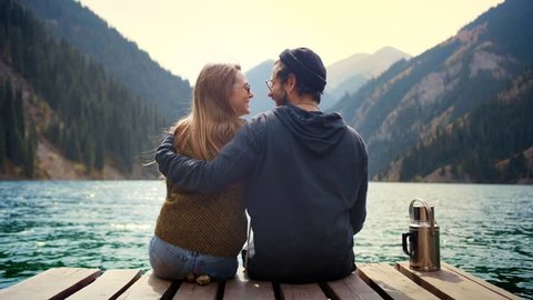 Beautiful real romantic couple of man and woman on first date in nature embraceing look to each other and kiss sitting on wooden bridge of mountains lake observing view and feel love calm happiness