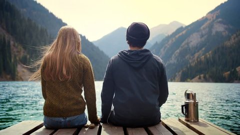 Beautiful real romantic couple of man and woman on first date in nature holding hands look to each other and kiss sitting on wooden bridge of mountains lake observing view and feel love calm happiness