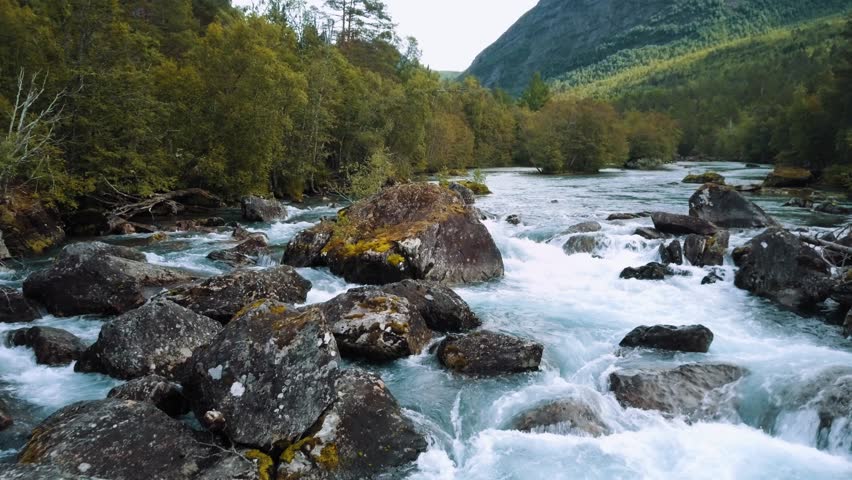 Drone panning above turquoise and white water river rapids with large boulders, orange trees and mountain ranges deep in a forest valley in Norway