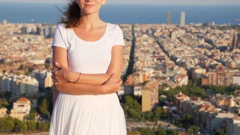 Portrait of young woman against urban landscape, slow motion shot, camera tilt up from white skirt to face. Lady stay folding arms, look straight in sunglasses. Blurred Barcelona cityscape seen back