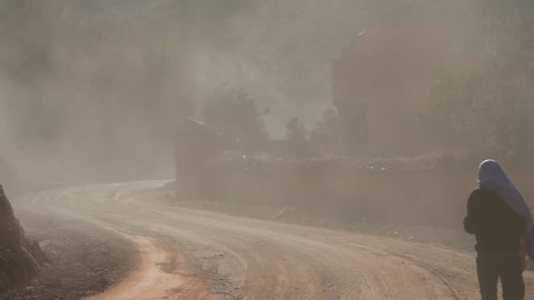 Lots of dust on a country road somewhere in Morocco, with a local man walking on the right side of the frame.