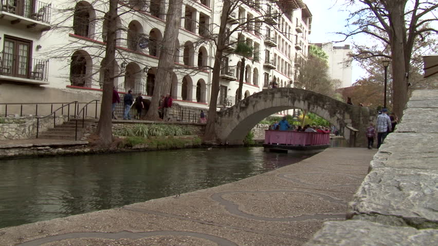 This is a side angle shot of the River Walk in San Antonio. Tourists and locals walk by enjoying the beautiful river.