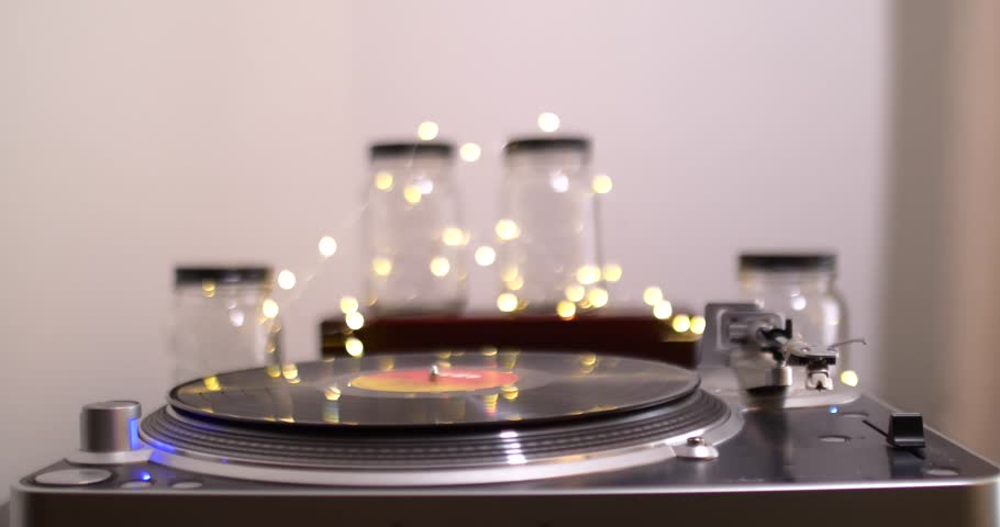 Male begins playing a vinyl record and setting the needle with lights and jars in background, lights dance on record player