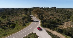 Aerial video of movement of red sport car on the twisting road among hills of Portugal, trees against a green grass, sunny weather, sunshine through trees, meadow, fields
