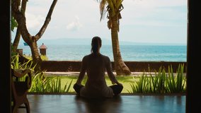 Silhouette of woman is practicing yoga meditation in lotus position, stretches hands up on the porch of bungalow on the ocean beach with cloudy sky, azure water, palm trees, green grass on island Bali
