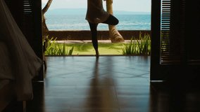 Silhouette of woman is practicing yoga meditation in tree position, stretches hands up on the ocean beach with beautiful view, green grass, nature sounds, birds singing, in Bali. Camera moves up