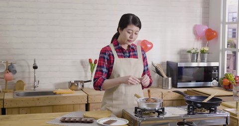 wife in apron standing in modern wooden kitchen breaking dark chocolate into pieces. asian female chef baker mixing sweet delicious organic melted cocoa cream in bowl in hot pot on stove stirring.