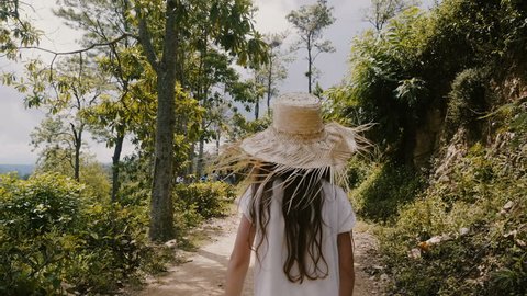 Camera follows happy little 5-7 years old girl child in big straw hat walk along sunny tropical forest road on vacation.