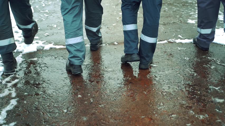 Four workers in a gray uniform with a horizontal white stripe on their pants are walking along the asphalt with wet snow in black boots. Close-up of legs. Royalty-Free Stock Footage #1025187557