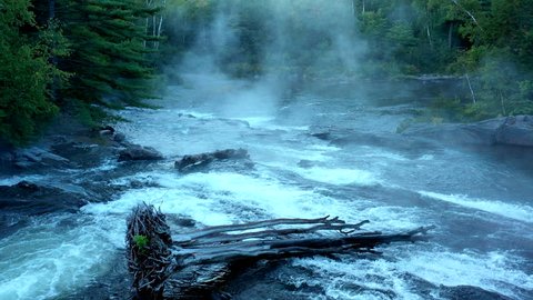 Aerial drone shot over the dark misty forest stream at Big Wilson Falls with logs in the waterfall at dusk in Maine.