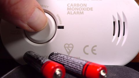 Close POV shot of a finger tip pressing the button to test the siren of  a carbon monoxide alarm, with red and green lights flashing and new batteries nearby, if needed.