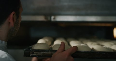 Slow motion close up of a baker pulling into the oven balls of dough covered with wheat flour ready for baking in bakery.