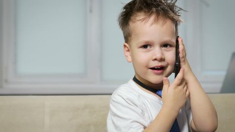 Close-up portrait of a little boy with tousled hair in a white shirt with a blue tie talking about something fun on a cell phone. Beautiful baby, fashionable kid in the office