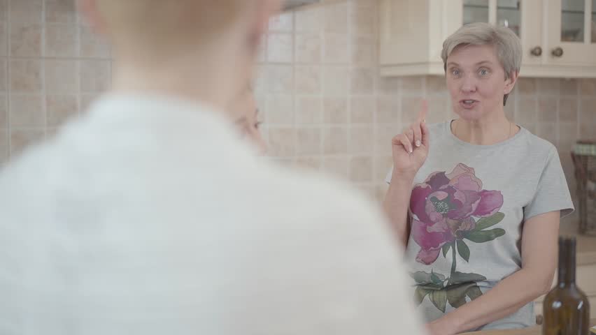 Group of three middle aged mature women communicating chatting in the kitchen at home with wine on the table. Shooting from behing one lady's back