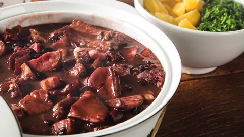 Delicious feijoada served in a white pot typical of Minas Gerais on a rustic wooden table accompanied by seasonings with smoke. Traditional Brazilian food.
