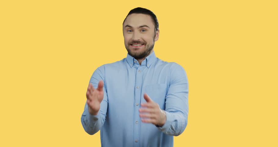 Handsome cheerful young man in the blue shirt standing on the yellow screen background, smiling archly while rubbing his hands.