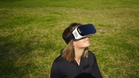 Young woman in sportswear using virtual reality headset in park. High angle view of sporty girl sitting on green lawn and enjoying virtual reality at sunny day, handheld shot. Technology concept