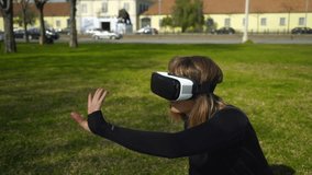 Girl using virtual reality headset in park. Vertical motion of young woman in sportswear sitting on lawn and using vr headset. Technology concept