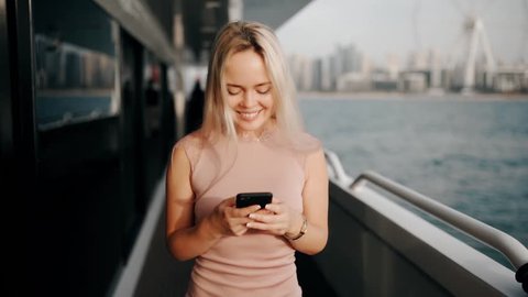 Beauty girl is walking on the yacht with a mobile phone in her hands and smiling. Dubai sea on the background.