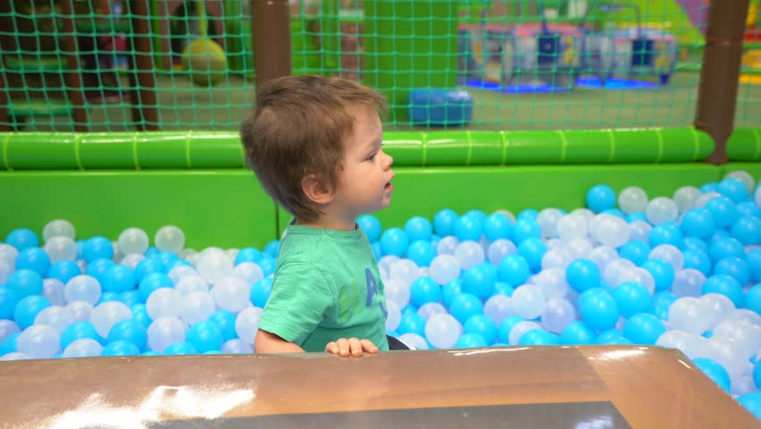 A boy of two or three years dressed in a turquoise t-shirt and dark blue pants alone stands in a dry pool with white and blue balls, then climbs out of it on a hill