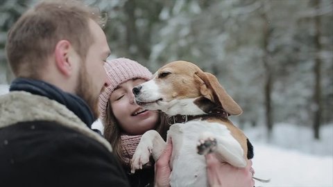 Cute young hipster couple having fun in winter park with their dog on a bright day and smiling. Man and woman playing with beagle. Slow motion,