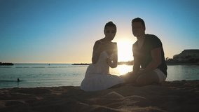A young couple sits on a sandy beach at sunset and communicates with friends via video call using an application on a smartphone