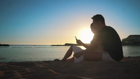 A young couple sits on a sandy beach at sunset and communicates with friends via video call using an application on a smartphone. Side view