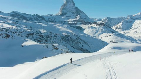 Blue Matterhorn Mountain in Winter Day and Hiker Man. Switzerland. Aerial View. Reveal Shot. Drone Flies Forward, Camera Tilts Up