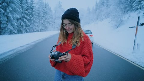 Young pretty female travel blogger or influencer, woman photographer in middle of beautiful dreamy winter snow covered road makes photos with camera, smiles and laughs, happy and authentic