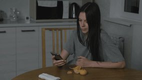 Woman watches a funny video at phone and eats a tangerine in the kitchen at night.