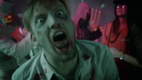 Halloween Costume Party: Brain Dead Bloody Zombie Turns Around and Attacks. In the Background Zombie, Death, Witch, Mummy and She Devil Have Fun in a Monster Party Decorated Room. Shot on 8K RED 