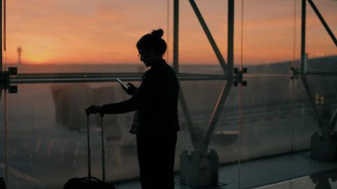 Blurred and unrecognizable young female traveler, leisure or business stands near glass window of big international airport hub with suitcase, scrolls through smartphone application,sunrise background