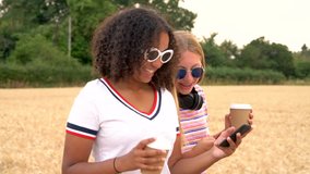 Slow motion tracking video clip of pretty blonde girl and mixed race teenager young women wearing sunglasses drinking coffee and using mobile cell smart phones for social media
