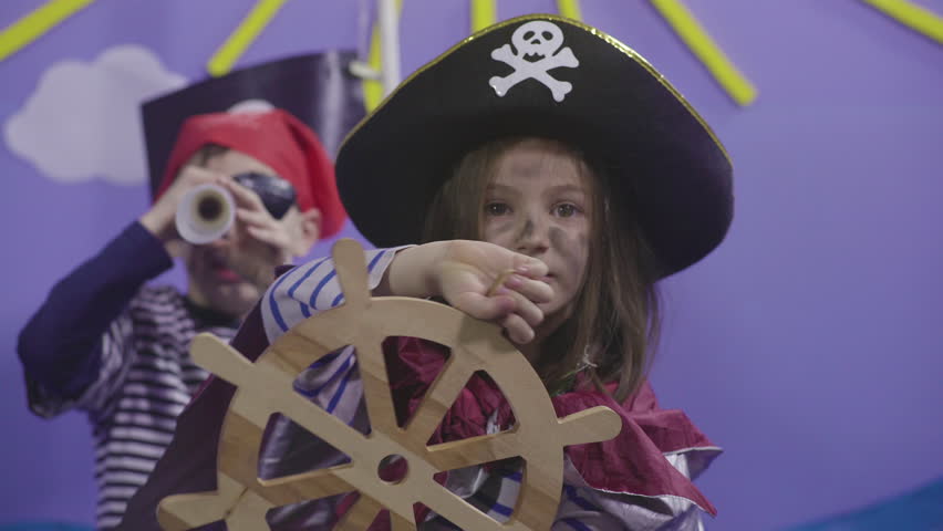 Two happy children acting pirates on sea scene in theatre stage, kids looking treasure, cute young girl captain holding and turning rudder, close up, in the background little boy using spyglass, 4k. Royalty-Free Stock Footage #1025328998