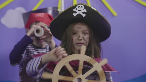Two happy children acting pirates on sea scene in theatre stage, kids looking treasure, cute young girl captain holding and turning rudder, close up, in the background little boy using spyglass, 4k.