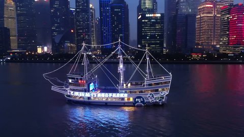 SHANGHAI - MARCH 21, 2018: Bright glowing cruise boat sail at Huangpu river against Lujiazui urban area, aerial orbiting shot. Night time, recreational vessel stylised on sailer illuminated by garland