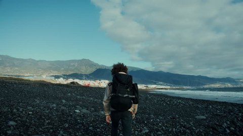 Epic and inspiring dreamy slow motion shot of camera follow adventurous young traveller, hitchhiker, camper or bikepacker walk towards incredible mountain landscape on beach, look back