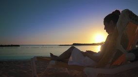 A young woman sits in a white dress on a sun lounger on a sandy beach at sunset and communicates with friends via video call using an application on her smartphone