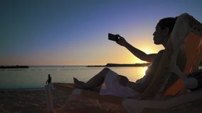 A young woman in a white dress sits on a sun lounger near the sea at sunset and communicates with friends via video call using the application on her smartphone