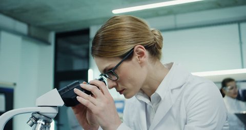 Close up of the beautiful young Caucasian woman in glasses working in the laboratory and looking in the microscope, then typing on the laptop computer. Camera zooming out.