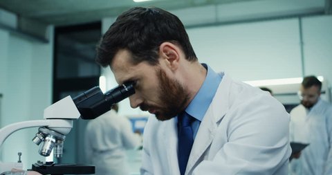 Close up of the attractive Caucasian man working in the laboratory and looking in the microscope, then typing on the laptop computer. Camera zooming out.