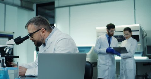 Close up of the Caucasian man in glasses, medical researcher, looking in the microscope while doing some analysis and typing them in the laptop computer at the laboratory.